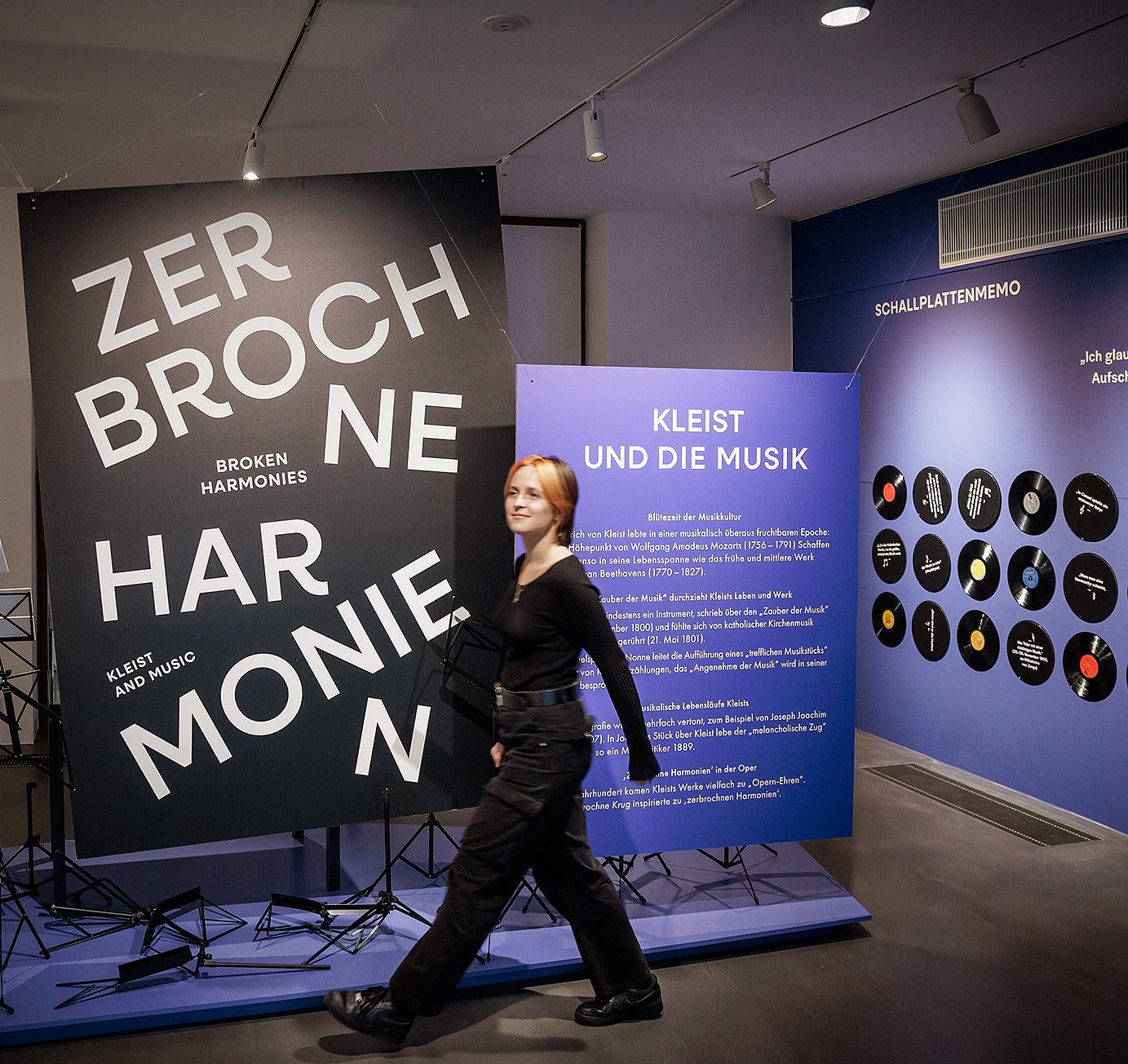 Blick in die Ausstellung ©Gordon Welters Im Vordergrund steht eine große, schwarze Tafel mit weißer, versetzt angeordneter Schrift: ‚ZERBROCHNE HARMONIEN‘. Links daneben sind Notenständer zu sehen. Im Hintergrund befindet sich eine blaue Wand mit einem Informationstext in weißer Schrift und der Überschrift ‚KLEIST UND DIE MUSIK‘. Rechts davon sind runde, schwarze Tafeln mit grafischen Elementen und kurzen Texten an der Wand angebracht. Der Raum ist hell ausgeleuchtet und wirkt modern gestaltet.